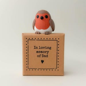 Könnte beinhalten: Eine dekorative Rotkehlchen-Figur mit roter Brust und braunen Flügeln sitzt auf einer braunen Schachtel. Die Schachtel hat einen schwarzen gepunkteten Rand und den Text "In loving memory of Dad" mit einem Herzsymbol.