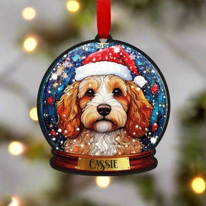 Könnte beinhalten: Ein Weihnachtsbaumschmuck aus Holz mit einem Hund, der einen Weihnachtsmann-Hut trägt, in einer Schneekugel. Der Hund ist ein weiß-brauner Cockapoo mit einer schwarzen Nase. Der Schmuck hat eine goldene Namensplakette mit der Aufschrift "Cassie".