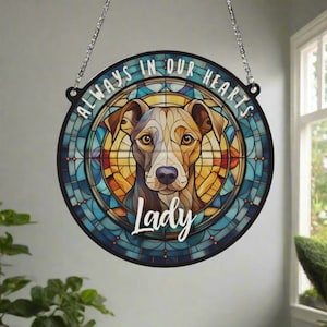 Pode incluir: Uma placa redonda de estilo vitral comemorativa com o rosto de um cão no centro. A placa é azul, amarela e laranja com uma borda preta. O texto "Always in our hearts" está escrito ao redor da parte superior da placa. O nome "Lady" está escrito abaixo do rosto do cão.