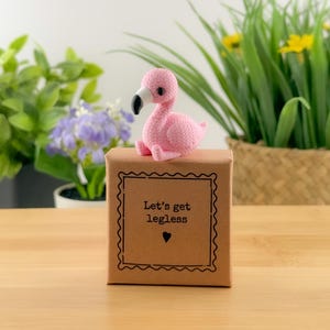 May include: A small pink flamingo figurine with a black and white beak sits atop a square brown cardboard box. The box features a decorative scalloped border and the printed black text "Let's get legless" above a small black heart icon.