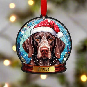 Peut inclure: Une boule de Noël ronde en bois noir avec un motif de boule à neige. À l'intérieur du globe se trouve un Braque allemand à poil court brun et blanc portant un bonnet de Père Noël rouge. Le nom du chien, "PENNY", est inscrit sur une plaque dorée au bas de l'ornement.