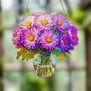 May include: A bouquet of purple and white flowers in a clear glass vase with the name "Emma" written on it. The vase is hanging from a silver chain.
