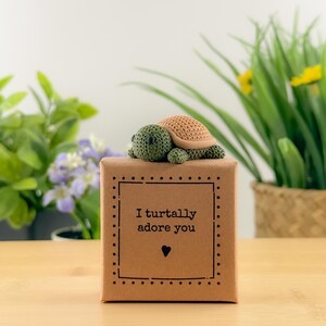May include: A crochet turtle figurine with a green head and feet, and a tan shell, sits on a brown gift box. The box has a black dotted border and the text "I turtally adore you" with a heart symbol.