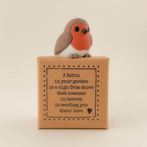 Könnte beinhalten: Eine handgefertigte Rotkehlchen-Figur mit roter Brust, braunen Flügeln und schwarzen Augen, die auf einer braunen Geschenkbox sitzt. Die Box enthält eine gerahmte Botschaft über Liebe und Erinnerung. Der Text lautet: „Ein Rotkehlchen in Ihrem Garten ist ein Zeichen von oben.“