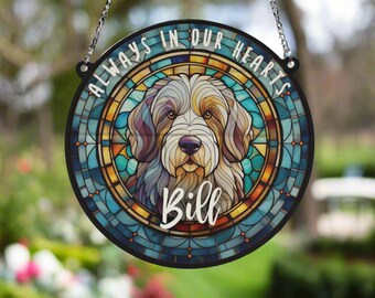 May include: A round, stained glass memorial plaque with a dog's face in the center. The plaque is blue, green, and yellow with a white border. The text "Always in our hearts" is written around the top of the plaque and the name "Bill" is written below the dog's face.