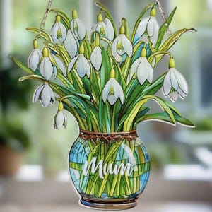 Può includere: Un ornamento appeso in stile vetrata con un mazzo di fiori bianchi di bucaneve in un vaso blu e verde con la scritta "Mamma" sul davanti.
