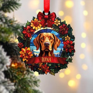 Könnte beinhalten: Ein runder Weihnachtsschmuck mit einem Vizsla-Hundeporträt in einem floralen Kranz. Der Schmuck hat ein rotes Band und eine Schleife oben. Der Name des Hundes, "LUNA", ist auf einem roten Banner unten zu sehen. Der Kranz enthält rote und gelbe Blumen.