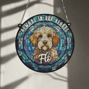 Pode incluir: Um ornamento memorial redondo de vidro colorido azul e branco com o rosto de um cão no centro. O cão é um doodle marrom claro e branco com o nome "Flo" escrito abaixo. O texto "Sempre em nossos corações" está escrito ao redor do ornamento.