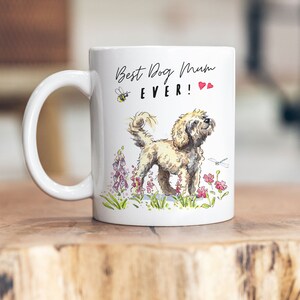 Könnte beinhalten: Weiße Keramiktasse mit einer Illustration eines braunen und weißen Hundes, rosa und lila Blumen, einer Biene und einer Libelle. Der Text auf der Tasse lautet "Best Dog Mum EVER!" mit zwei roten Herzen.