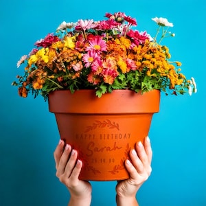May include: A terracotta flower pot filled with a vibrant arrangement of pink, yellow, orange, and white chrysanthemums. The pot features an engraved wreath design with the text "HAPPY BIRTHDAY Sarah 25.10.80" centred on the front surface.