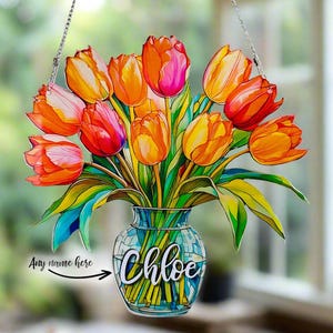 Op de afbeelding: Een hangende decoratie in de stijl van glas-in-lood met een boeket oranje, gele en roze tulpen in een blauwe en groene mozaïekvaas. De vaas heeft de naam "Chloe" erop.