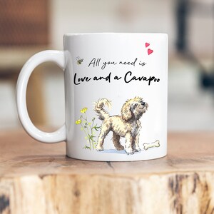 Könnte beinhalten: Weiße Keramiktasse mit einer Cartoon-Illustration eines Cavapoo-Hundes, gelben Blumen und einer Biene. Der Text auf der Tasse lautet "All you need is Love and a Cavapoo".