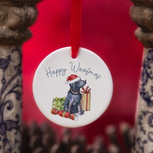May include: White ceramic Christmas bauble featuring a black dog wearing a Santa hat and holding a gift. The bauble reads "Happy Woofmas".
