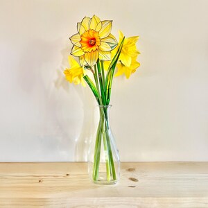 May include: A bouquet of yellow daffodils in a clear glass vase. The daffodils have long green stems and are arranged in a vase on a light wood surface.