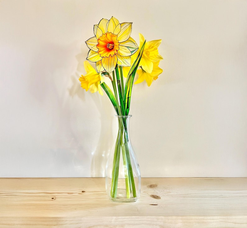 May include: A bouquet of yellow daffodils in a clear glass vase. The daffodils have long green stems and are arranged in a vase on a light wood surface.