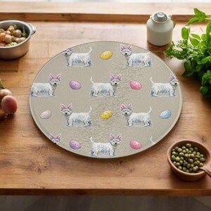 Könnte beinhalten: Ein rundes Schneidebrett aus klarem Glas mit einem sich wiederholenden Muster aus weißen Hunden mit rosa Hasenohren und bunten Ostereiern. Das Brett liegt auf einer Holzoberfläche, mit anderen Küchenartikeln im Hintergrund.