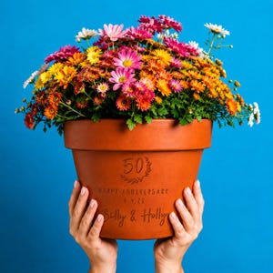 Può includere: Un vaso da fiori in terracotta riempito con una vivace composizione di crisantemi rosa, arancioni, gialli e bianchi. Il vaso è inciso con il numero 50, il testo HAPPY ANNIVERSARY 6.4.22 e i nomi Billy & Holly in un carattere corsivo.