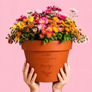 Può includere: Un vaso da fiori in terracotta riempito con una vivace composizione di crisantemi rosa, arancioni, gialli e bianchi. Il vaso è inciso con il testo HAPPY RETIREMENT, la data 1.5.22 e il nome Marilyn, incorniciato da motivi a foglie decorativi.