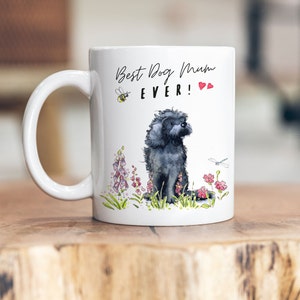 Può includere: Tazza in ceramica bianca con un'illustrazione in bianco e nero di un cane seduto in un campo di fiori. Il testo "Best Dog Mum EVER!" è stampato sulla tazza.