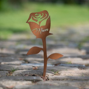 May include: A rusty metal rose with a cut-out design and the word "Mum" inscribed on the rose. The rose is standing upright on a bed of grey stones.