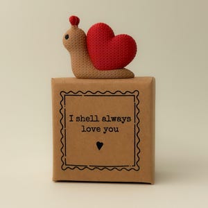 May include: A knitted snail ornament with a red heart-shaped shell rests on a brown gift box. The box is inscribed with the words "I shell always love you" within a decorative border, and a heart symbol.