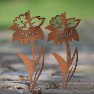 May include: Two metal flower garden stakes with a rust finish. One stake reads "Mum" and the other reads "Dad". The flower petals have a cutout design with green accents. The stakes are placed on a stone surface.