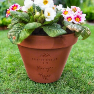 K&ouml;nnte beinhalten: Ein Terrakotta-Blumentopf mit wei&szlig;en und rosafarbenen Primeln. Der Topf ist mit einer Gravur versehen, die den Text "HAPPY RETIREMENT 1.5.22" und den Namen "Marilyn" zwischen zwei kleinen Blattzweig-Illustrationen zeigt.