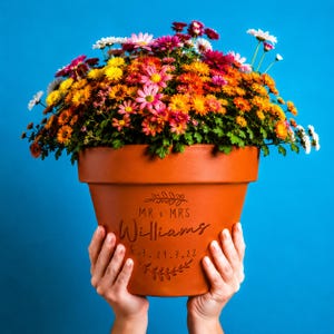 Può includere: Un vaso da fiori in terracotta riempito con una densa composizione di crisantemi dai colori vivaci giallo, arancione, rosa e bianco. Il vaso presenta un'incisione con il testo MR & MRS Williams Est. 29.3.22, incorniciato da motivi a foglia.