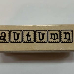 May include: A rectangular wooden stamp with the word "AUTUMN" printed in black, block letters. Each letter is enclosed in a square with a dotted border. The stamp is light brown.