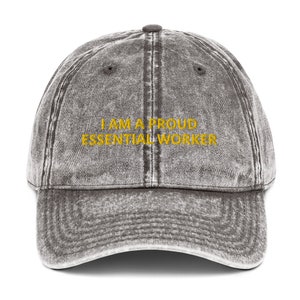 May include: A grey vintage baseball cap with the text "I AM A PROUD ESSENTIAL WORKER" embroidered in yellow.