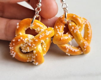 May include: A pair of earrings featuring miniature pretzel charms with a salt coating. The earrings are made of silver and have a simple hook design.