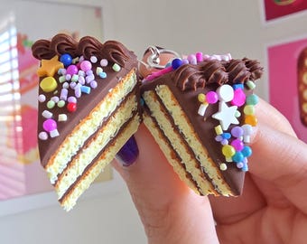 May include: Two miniature cake slice charms. Each slice has a chocolate frosting top with decorative sprinkles and a star. The cake layers are a light yellow color. The charms are held up by a silver ring.