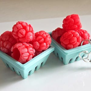May include: Earrings with miniature light blue baskets filled with bright red raspberries. The earrings have silver-coloured hooks. The raspberries appear to be made of a solid material. The background is white. The earrings are a fun, quirky accessory.