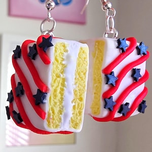 May include: A pair of dangle earrings featuring miniature cake slices with white frosting, yellow sponge cake layers, red icing swirls, and small dark blue star-shaped decorations. The earrings are attached to silver-toned metal hooks for hanging.