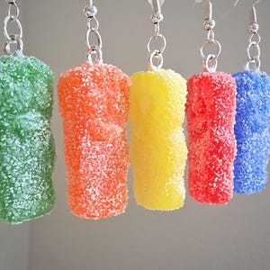 May include: A set of five dangling earrings shaped like gummy bears in green, orange, yellow, red, and blue. Each bear has a textured, sugar-coated appearance and is attached to a silver-toned metal jump ring and a standard earring hook.