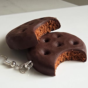 May include: Chocolate cookie earrings with a bite taken out of one. The dark brown cookies have a textured, light brown filling and small holes on the top. The earrings have silver-colored hooks and a small bead.