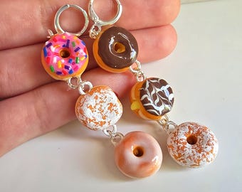 May include: A pair of dangle earrings featuring miniature donut charms. The donuts have various toppings, including sprinkles, chocolate frosting, and powdered sugar. The earrings are connected with silver-colored metal links and have hoop closures.