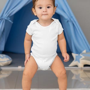 May include: A baby in a white onesie stands on a play mat with a road design. The baby is looking at the camera and has a serious expression on their face.