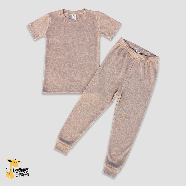 May include: A two-piece set of pajamas for children, featuring a short-sleeved t-shirt and long pants. Both pieces are a light brown color with a subtle heather texture.