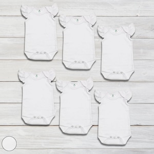 May include: Six white baby onesies with ruffled sleeves. The onesies are laid out on a white wooden surface.