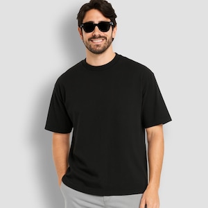May include: A black crew neck t-shirt. The short-sleeved shirt is worn by a person wearing black sunglasses. The person is smiling and has a beard. The background is a solid light gray.