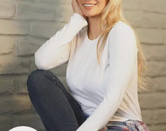May include: A woman wearing a white long-sleeved shirt, blue jeans, and a brown leather baseball cap. She is sitting with her hand on her cheek and smiling. A plaid shirt is tied around her waist.