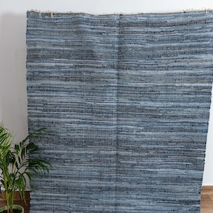Puede incluir: Una alfombra rectangular grande con apariencia de mezclilla azul. La alfombra está hecha de tiras de mezclilla tejidas en diferentes tonos de azul, creando una superficie texturizada. Los bordes tienen flecos. Una planta en maceta es visible en la esquina.