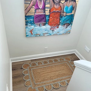 May include: A brown and beige jute rug with a scalloped edge and a blue trim. The rug is on a wooden floor in a hallway with a white wall and a painting of three women in swimsuits.