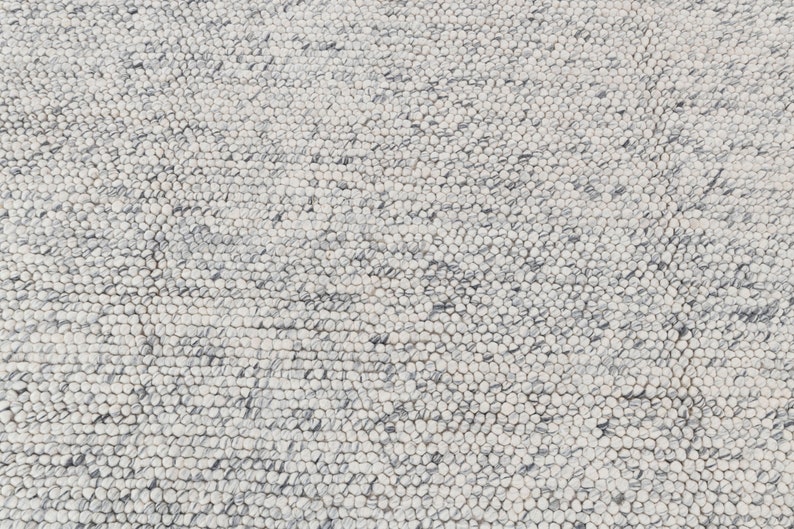 May include: Close-up of a white and gray textured rug with a circular pattern. The rug is made of small, round, woven fibers.