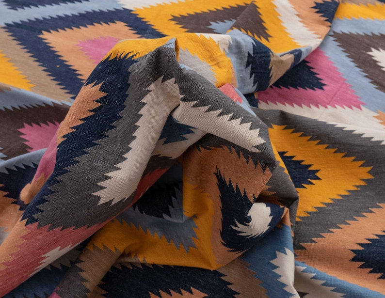 May include: A colorful woven fabric with a geometric pattern in shades of blue, yellow, pink, and brown. The fabric is folded and draped, creating a textured and abstract design.
