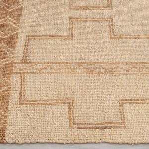 May include: A natural fiber rug with a beige background and a brown geometric pattern. The rug has a woven texture and a fringed edge.