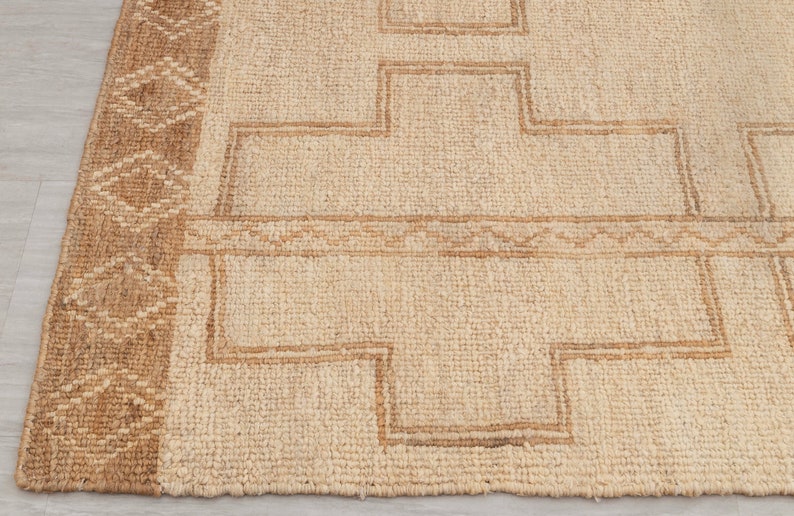 May include: A natural fiber rug with a beige background and a brown geometric pattern. The rug has a woven texture and a fringed edge.