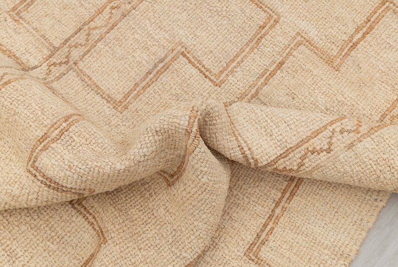 May include: A cream-colored rug with a geometric pattern woven in a darker beige color. The rug is folded in half, showing the texture of the woven fibers.