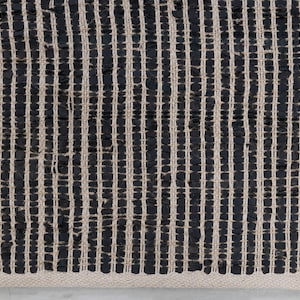 Puede incluir: Alfombra de yute negra y natural con un patr&oacute;n de rayas tejido. La alfombra tiene un borde con flecos.